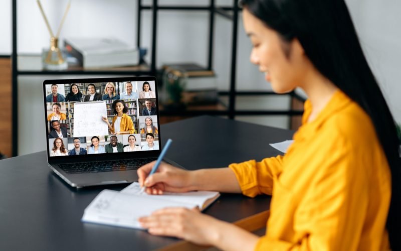 Online education. Smart japanese student girl gains knowledge watching lesson online. Young woman takes notes during webinar, listening lecture on video call. Female teacher and students on a screen